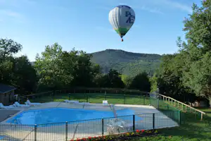 Schwimmbecken auf dem Campingplatz mit Zaun, dahinter Bäume und ein Hügel, am Himmel ein Heißluftballon.