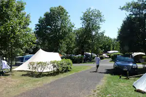 Weißes Zelt auf einem Campingplatz neben einem gepflasterten Weg, daneben geparkte Autos und Bäume im Hintergrund.