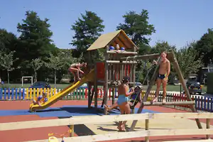 Spielplatz am Campingplatz mit Holzspielgerüst, Rutsche und Kindern am Planschbecken, im Hintergrund Bäume und Fahrzeuge.