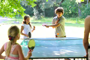 Tischtennis am Campingplatz: Kinder spielen an einer blauen Tischtennisplatte im Grünen unter Bäumen.