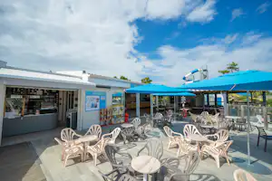 Terrasse am Campingplatz mit weißen Kunststoffstühlen, Tischen und blauem Sonnenschirm vor einem Verkaufsbereich.