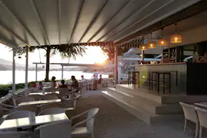 Terrasse am Campingplatz mit weißen Stühlen, Tisch, Barbereich und Blick auf Wasser und Küste bei Sonnenuntergang.