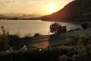 Sonnenuntergang an der Bucht: ruhiges Wasser, Boote vor der Küste und bewaldete Hügel im Hintergrund.