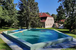 Großes Camping-Schwimmbecken im Garten mit Rasen, Bäumen und einem weißen Gebäude mit rotem Dach im Hintergrund.
