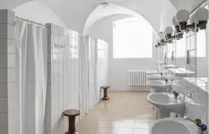 Modernes Badezimmer mit mehreren Waschbecken, Armaturen und weißen Fliesen, Duschkabinen mit Vorhängen und Rundbogenfenstern.