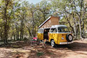 Gelbes Wohnmobil mit aufgestelltem Dachzelt im Wald, daneben Campingstühle und ein Tisch auf dem Stellplatz.