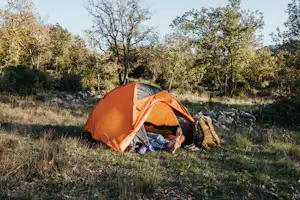 Orangefarbenes Zelt auf einer Wiese am Campingplatz, mit Rucksack und Schlafsack vor einem Steinwall und Bäumen im Hintergrund.