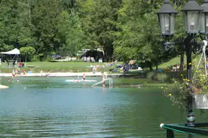 Badebereich am Camping-See mit Menschen im Wasser, Liegen und Bäumen im Hintergrund.