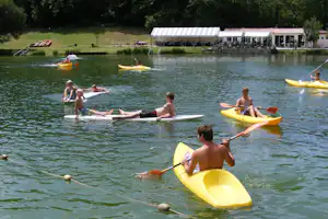 Mehrere Personen paddeln mit gelben und roten Kajaks auf dem Campingsee, im Hintergrund Ufer mit Terrasse und Gebäuden.