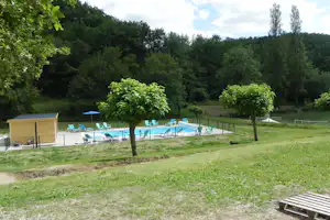 Schwimmbecken mit blauen Liegen und Sonnenschirmen, umgeben von Rasen, Bäumen und einem kleinen Holzhaus am Campingplatz.