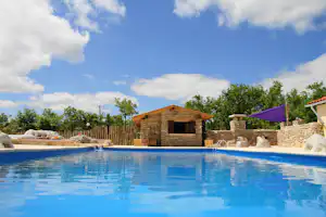 Blauer Camping-Pool mit Steinmauer, Holz-Pavillon und lila Sonnenschirm im Hintergrund, umgeben von Bäumen und Zaun.