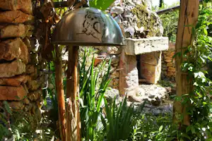 Gartenbrunnen mit Metallkugel und Steinmauer, umgeben von Pflanzen und Gras auf einem Campingplatz.