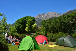 Grüne und rote Campingzelte auf einer Wiese, daneben Campingstühle und Tisch, im Hintergrund Bäume und Berge.
