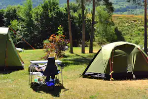 Zwei Campingzelte auf einer Wiese zwischen hohen Bäumen, im Hintergrund Berge und ein weiterer Zeltplatz.