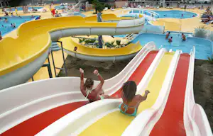 Gelbe und rote Wasserrutschen am Pool eines Campingplatzes, daneben weitere Becken und Liegen.