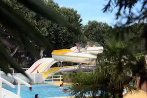 Gelbe Wasserrutsche über dem Pool, umgeben von Palmen und Bäumen, im Hintergrund ein Gebäude am Campingplatz.