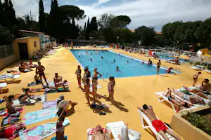 Großes Camping-Schwimmbecken mit Sonnendeck, Liegen und vielen Badegästen, umgeben von Bäumen und einem Zaun.