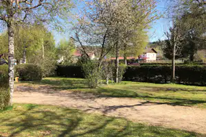 Grüner Campingplatz mit Rasen, Bäumen und Hecken, im Hintergrund Gebäude und ein Weg.