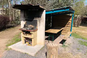 Feuerstelle mit Grill im Campingbereich, überdachter Holzunterstand, Holztisch und Stühle im Garten zwischen Bäumen.
