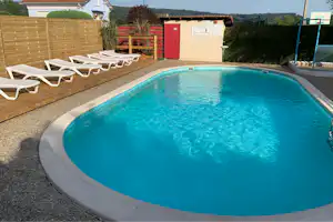 Rundes Camping-Pool mit blauem Wasser, umgeben von Kiesweg, Holzsichtschutz und mehreren weißen Liegen.