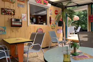 Terrasse am Campingplatz mit runden und rechteckigen Tischen, Stühlen und Deko an einer Holzfassade.