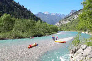 Flussufer mit mehreren Kajaks am Strand, Menschen im Wasser und Berge im Hintergrund, umgeben von grünen Bäumen.