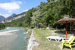 Campingplatz am Fluss mit Kiesufer, Liegestühlen und Sonnenschirm unter Bäumen, im Hintergrund Berge und grüner Hang.
