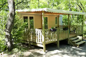 Bungalow mit Holzveranda und Stufen, davor ein Tisch und Stühle, umgeben von Bäumen im grünen Campingbereich.
