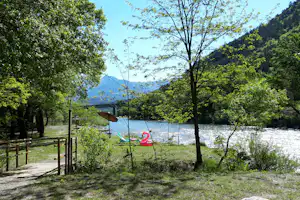 Campingplatz am See mit Uferweg, Bäumen und Blick auf die Berge in der Ferne.