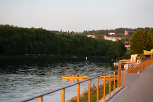 Blick vom Uferweg auf den See mit Steg und Geländer, im Hintergrund Häuser und Bäume.