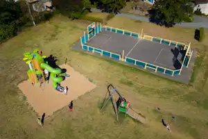 Tennisplatz auf dem Campinggelände mit Umrandung, daneben Rasenflächen und Wege, im Hintergrund Gebäude und Bäume.