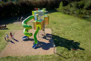 Spielplatz am Campingplatz mit grünem Rutschenturm, Sandfläche und Bäumen im Hintergrund.