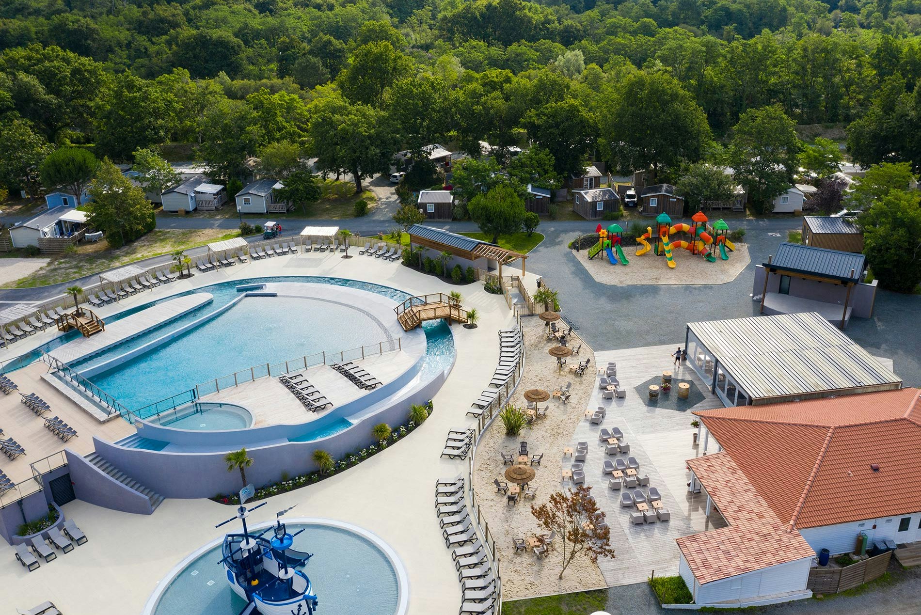 Luftaufnahme von Campinganlage mit großem Schwimmbecken, Liegen, Spielplatz und mehreren kleinen Ferienhäusern im Grünen.