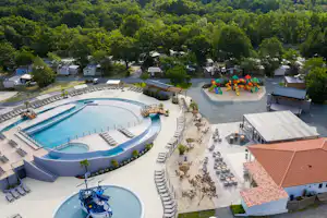 Luftaufnahme von Campinganlage mit großem Schwimmbecken, Liegen, Spielplatz und mehreren kleinen Ferienhäusern im Grünen.