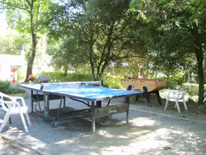 Camping-Tisch mit blauer Plane, daneben weiße Plastikstühle, im Schatten von Bäumen auf einem gepflasterten Stellplatz.