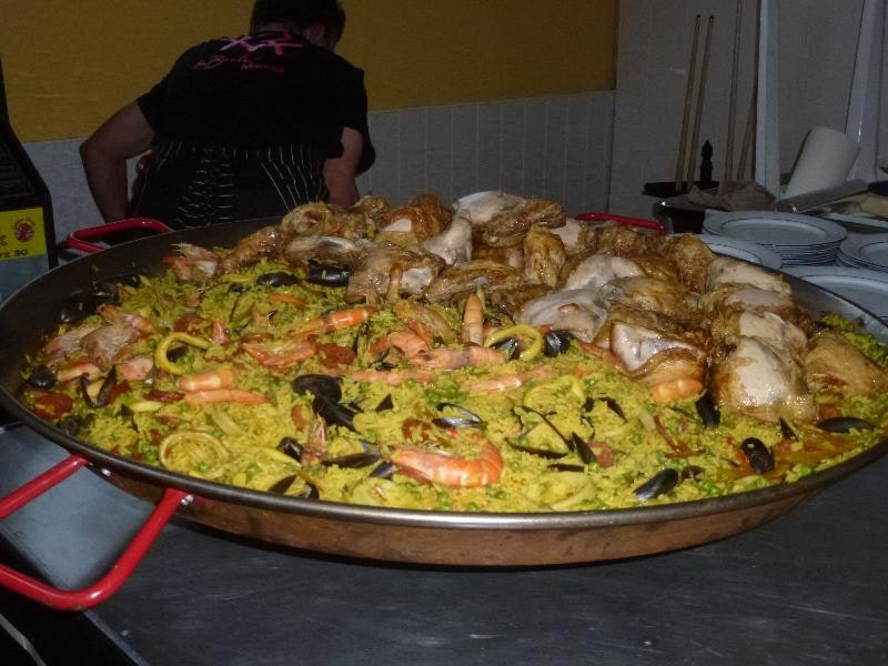 Große Paella-Pfanne mit Meeresfrüchten und Gemüse auf einem Camping-Tisch, daneben Teller und Besteck.