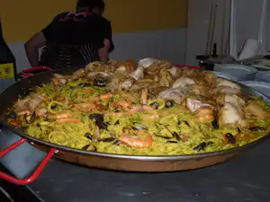 Große Paella-Pfanne mit Meeresfrüchten und Gemüse auf einem Camping-Tisch, daneben Teller und Besteck.
