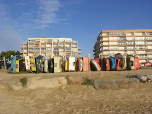 Reihe von Strandduschen und Bungalows am Campingplatz, dahinter mehrstöckige Gebäude und ein Sandweg.