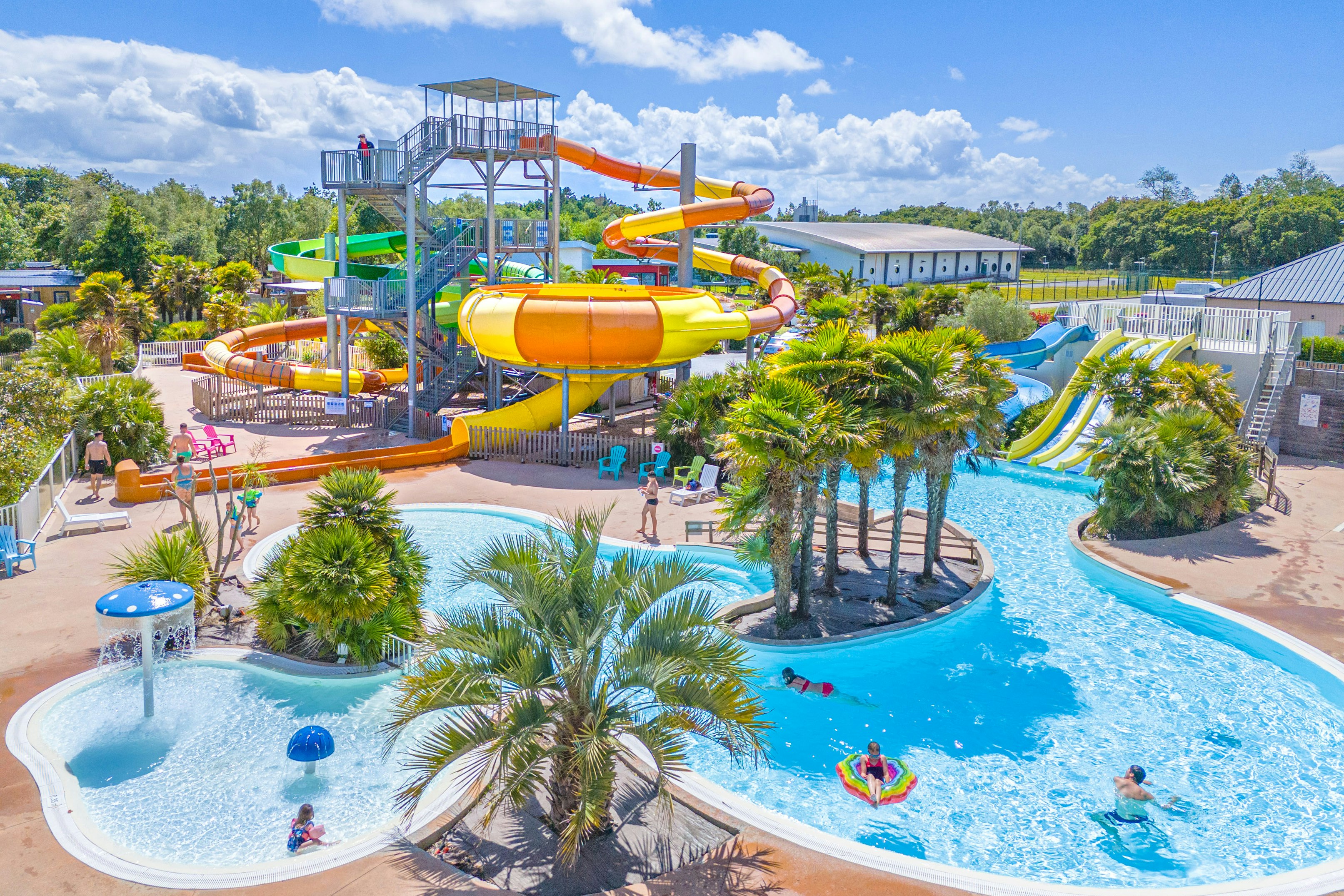 Großer Camping-Pool mit Wasserrutsche, Kinderbereich und Liegen, umgeben von Palmen und Bäumen, daneben Gebäude und Wege.