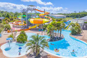 Großer Camping-Pool mit Wasserrutsche, Kinderbereich und Liegen, umgeben von Palmen und Bäumen, daneben Gebäude und Wege.