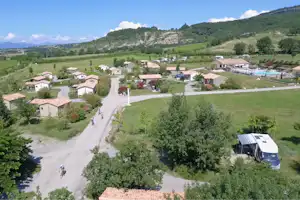 Campinganlage mit mehreren Mobilheimen, gepflasterten Wegen, Terrasse und Poolbereich, umgeben von grünen Hügeln und Bäumen.