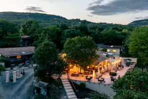 Blick auf den Campingplatz mit beleuchteten Terrassen, Sitzbereichen und Wegen zwischen Bäumen, im Hintergrund Hügel und Gebäude.