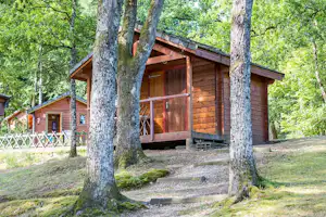 Holzhütte mit Veranda im Wald, Holzfassade und Treppe, umgeben von Bäumen und Kiesweg am Campingplatz.