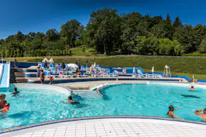 Schwimmbad mit blauem Becken, Liegestühlen und Sonnenschirmen auf der Terrasse, im Hintergrund grüner Hang mit Bäumen.