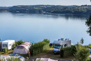 Campingplatz am See: Zelte und Wohnwagen auf dem Stellplatz, Hecken und Blick auf ruhiges Wasser und Hügel im Hintergrund.