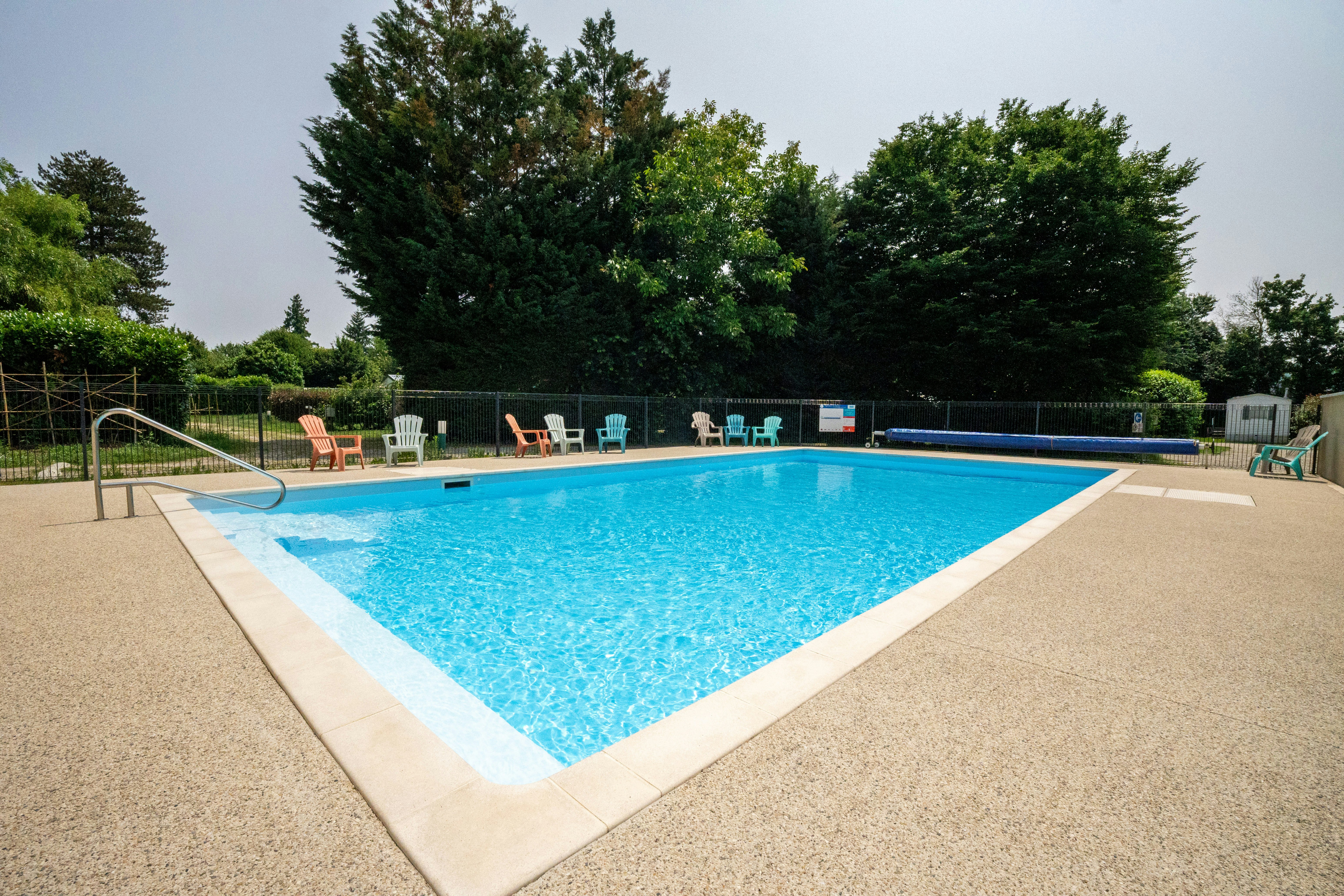 Schwimmbecken auf dem Campingplatz mit Liegen am Beckenrand, umgeben von Bäumen und Hecken.