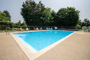 Schwimmbecken auf dem Campingplatz mit Liegen am Beckenrand, umgeben von Bäumen und Hecken.