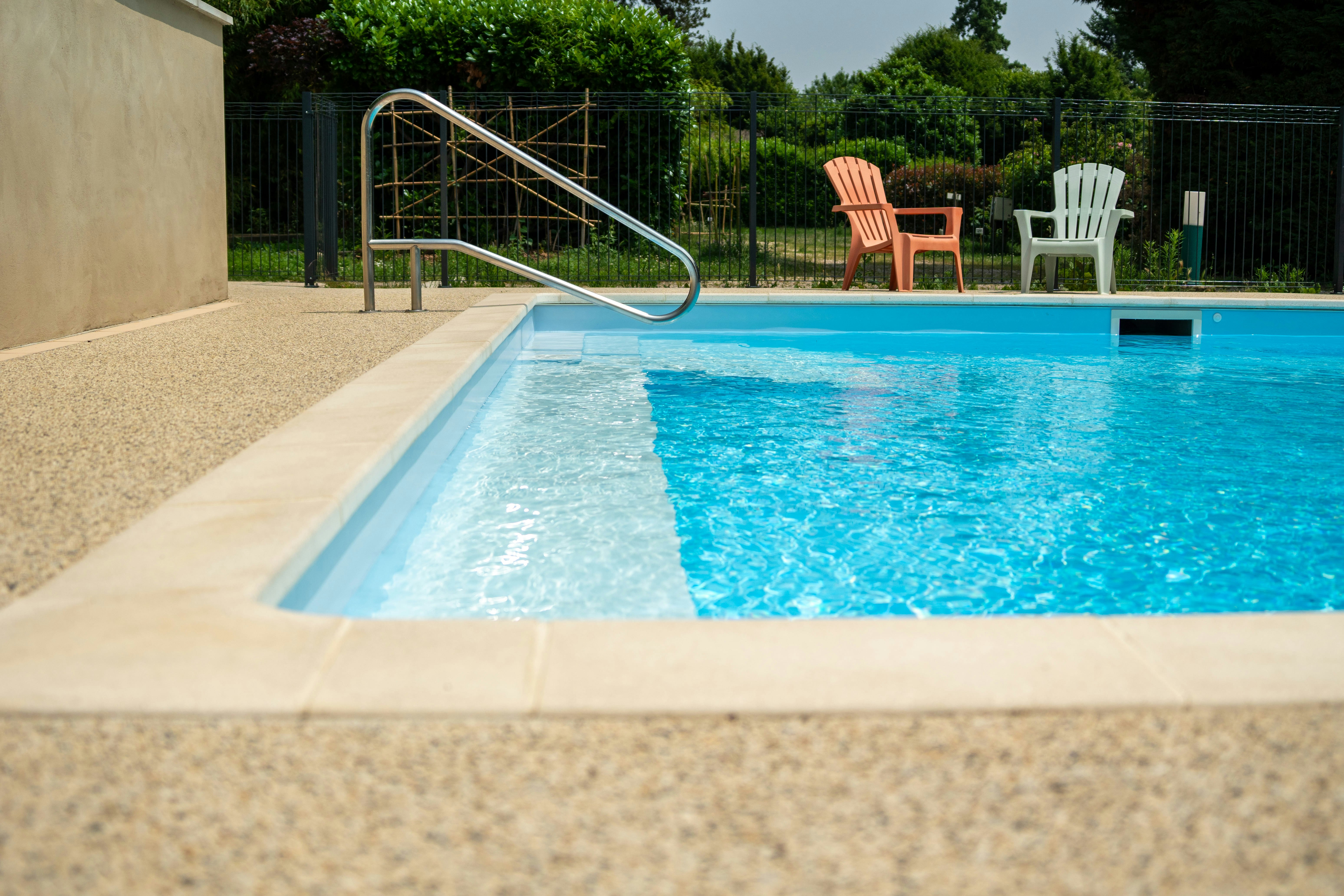 Blauer Camping-Pool mit Edelstahl-Leiter, beigen Fliesen und zwei weißen Liegestühlen neben dem Zaun.
