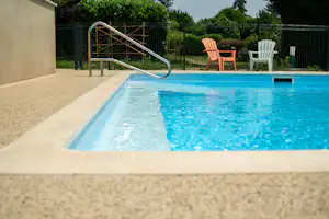 Blauer Camping-Pool mit Edelstahl-Leiter, beigen Fliesen und zwei weißen Liegestühlen neben dem Zaun.