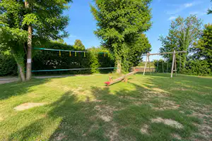 Spielplatz auf dem Campingplatz: grüner Rasen, Schaukel und Klettergerüst im eingezäunten Bereich mit Bäumen.